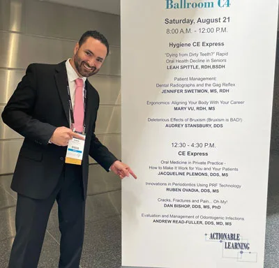 Image of Dr. Ovadia standing in front of a convention sign pointing at his name on the board of lecturers.